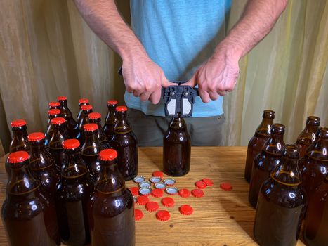 Craft beer brewing at home, man closes brown glass beer bottles with plastic capper on wooden table with red crown caps. Horizontal stock image