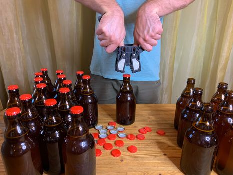 Craft beer brewing at home, man closes brown glass beer bottles with plastic capper on wooden table with red crown caps. Horizontal stock image