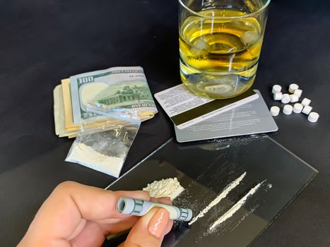 A female's hand holding rolled 100 us dollars banknote near two lines of cocaine. Plastic bag with illegal drugs, tablets, money and a glass of whiskey on background. Drug addiction. Stock image.