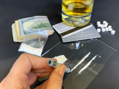A female's hand holding rolled 100 us dollars banknote near two lines of cocaine. Plastic bag with illegal drugs, tablets, money and a glass of whiskey on background. Drug addiction. Stock image.