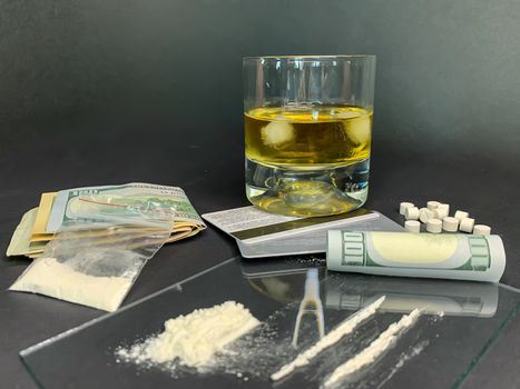Rolled 100 us dollars, 2 lines of cocaine, a glass of whiskey, plastic bag with illegal drugs and credit cards on a gray background. Drug addiction. Stock image.