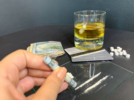 A female's hand holding rolled 100 us dollars banknote near two lines of cocaine. Plastic bag with illegal drugs, tablets, money and a glass of whiskey on background. Drug addiction. Stock image.