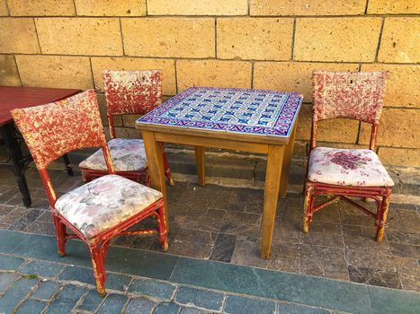 Antique table with blue mosaic tiles and three red old chairs near stone wall