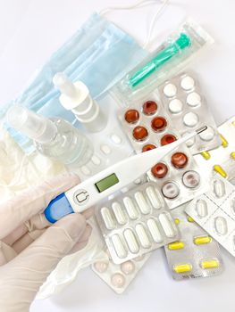 Digital thermometer for measuring body temperature in hand in glove of a woman on white background with many pills and medicines. Vertical image