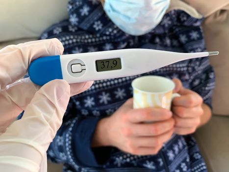 Hand in medical glove holds a digital thermometer. Sick male man patient with medical face mask with cup of hot tea on a background