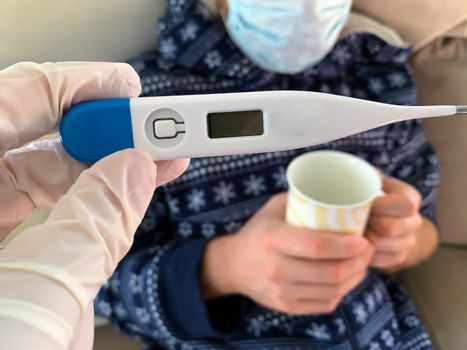 Hand in medical glove holds a digital thermometer. Sick male man patient with medical face mask with cup of hot tea on a background