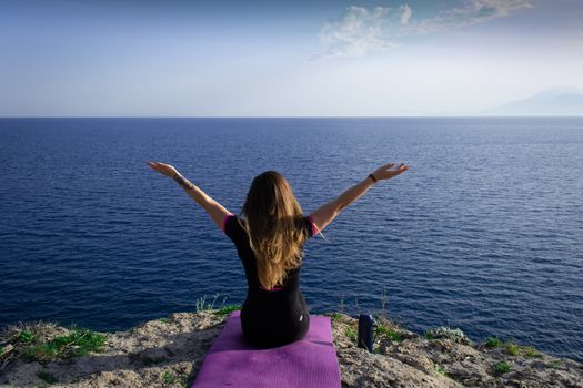 Beautiful young happy lonely girl sitting on rock and watching blue Mediterranean sea and relaxing