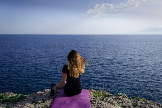 Beautiful young happy lonely girl sitting on rock and watching blue Mediterranean sea and relaxing