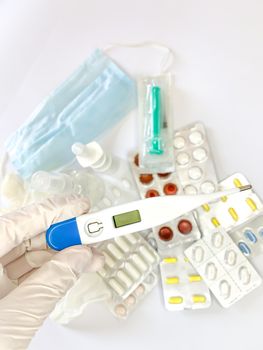 Digital thermometer for measuring body temperature in hand in glove of a woman on white background with many pills and medicines. Vertical image