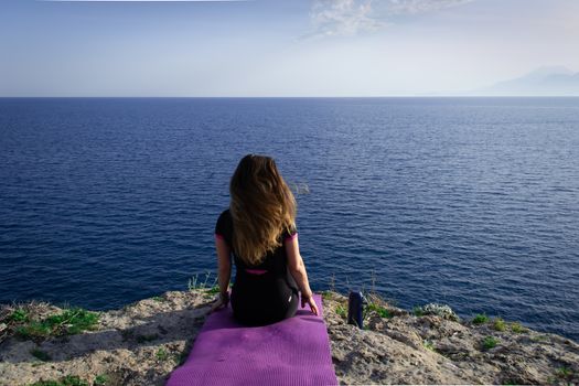 Beautiful young happy lonely girl sitting on rock and watching blue Mediterranean sea and relaxing