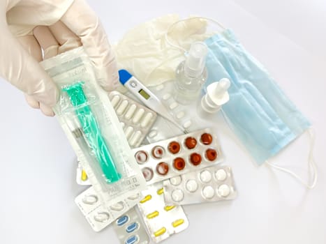 Syringe in hand in glove of a woman on white background with many pills and medicines. Horizontal image