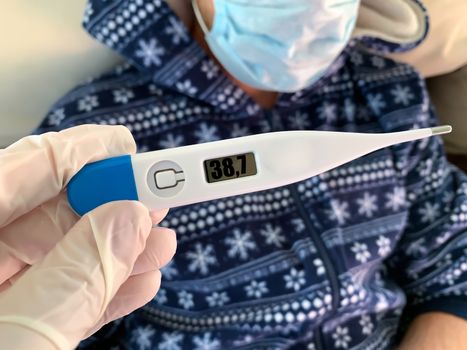 Hand in medical glove holds a digital thermometer. Sick male man patient in blue with medical face mask on a background