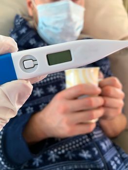 Hand in medical glove holds a digital thermometer. Sick male man patient with medical face mask with cup of hot tea on a background