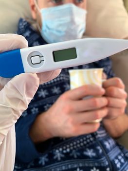 Hand in medical glove holds a digital thermometer. Sick male man patient with medical face mask with cup of hot tea on a background