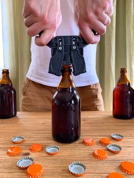 Craft beer brewing at home, man closes brown glass beer bottles with plastic capper on wooden table with orange crown caps. Vertical image