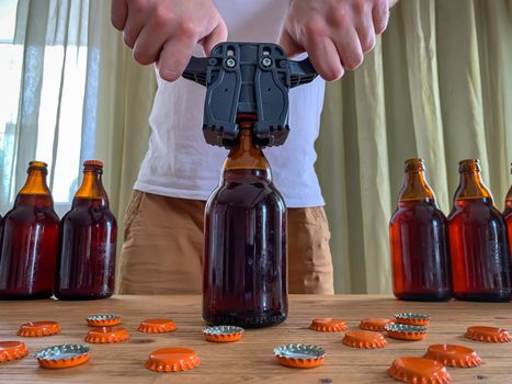 Craft beer brewing at home, man closes brown glass beer bottles with plastic capper on wooden table with orange crown caps. Horizontal image