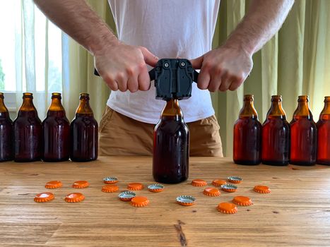 Craft beer brewing at home, man closes brown glass beer bottles with plastic capper on wooden table with orange crown caps. Horizontal image