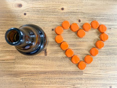An orange love heart made from beer bottle tops lids and a brown glass beer bottle on a rustic wooden table. Beer drinkers and lovers Valentine's day concept top view horizontal image