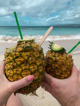 Two caucasian hands holding a pina colada cocktail in pineapple with green straws on a tropical beach in Dominican Republic. Vertical image