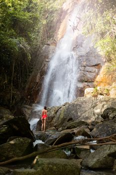 Beautiful young slim blonde woman with curly hair, wearing in red swimsuit is enjoy in lagoon of huge tropical waterfall in jungle. Travel concept. Sexy woman wearing red swimsuit in the jungle