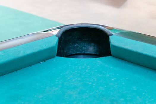 The pocket or hole of a billiard table that looks a little used and is covered with green billiard cloth. The pocket is protected with plastic and in the middle of two bands.