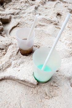 Two plastic cups standing in the sand on a beach and filled with a rest of drinks, which looks like cola and the other drink is turquoise. In both cups there is a white straw.
