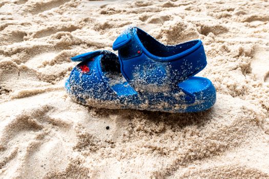 A blue children's sandal that stands in the sand on a beach and is soiled with sand by moisture. The velcro buckles are partially opened.