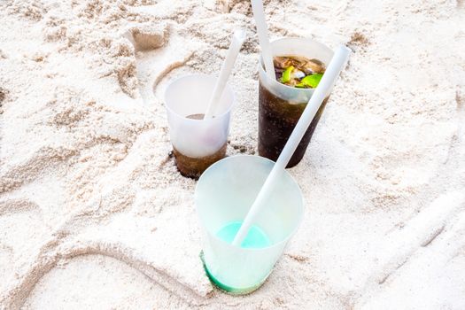 Three plastic cups stand in the sand, which are still partly filled with drinks. Two cups are filled with cola and limes, one cup with a turquoise drink. In all cups there is a white straw.