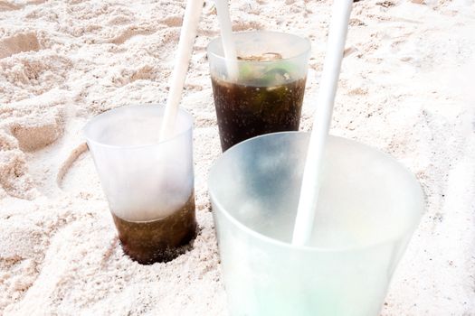 Three transparent plastic cups with white straws on the beach, partly filled with a dark drink, partly already drunk from it.