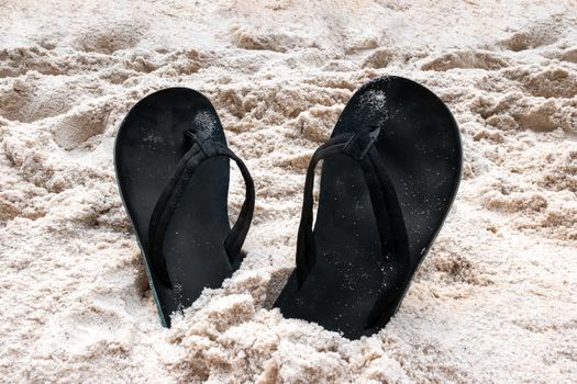 Two black flip-flop flip-flops with turquoise soles, stuck in the sand with their heels and slightly sanded.