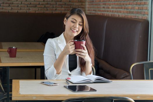 Asian businesswoman sitting and drinking coffee in a coffee shop Relax in the morning. On the table are books, tablets and mobile phones.
