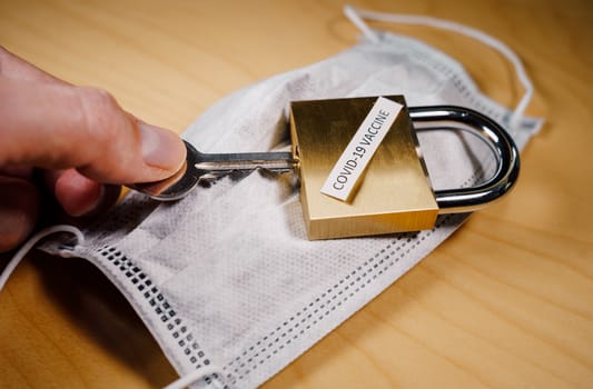 Hand opening a padlock over a face mask, symbolizing the discovery of the COVID-19 vaccine and the world lockdown end.