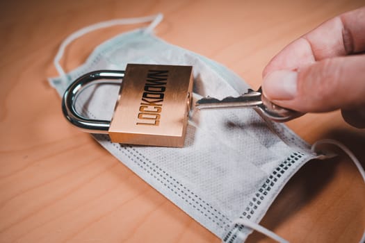 Hand opening a padlock over a face mask, symbolizing the discovery of the COVID-19 vaccine and the world lockdown end.