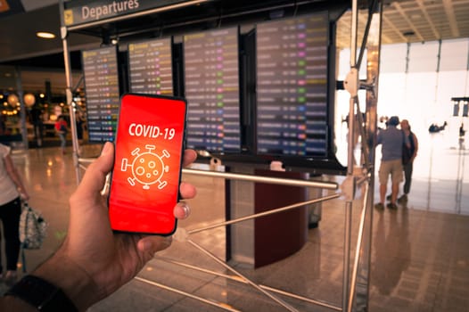 Hand holding a smartphone with Covid-19 message on screen and airport timetables as background. Symbolizing the global spread of the coronavirus through global air traffic