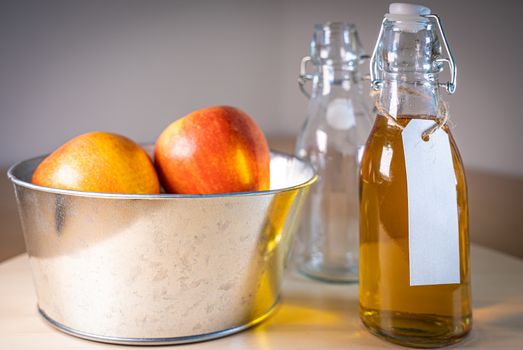 Close up view of glass bottle with a blank tag of apple juice or cider and apples in basket on wooden tabletop
