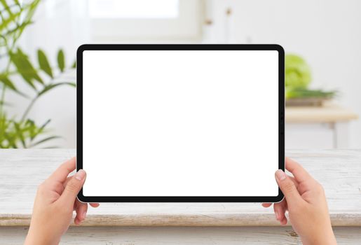 Isolated human left hand holding black tablet media device with white empty screen mockup and wooden table in kitchen