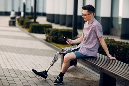 Disabled young man with foot prosthesis sitting and hold mobile phone outdoor