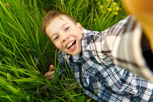 Happy smiling boy making self portrait on smartphone in meadow. young boy making selfie on smartphone laying in green grass with yellow flowers