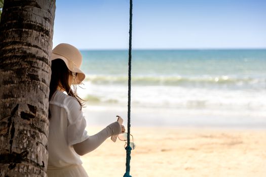 Summer concept. Asian woman tourist with white dress and hat relax on swing on the beach and sea.