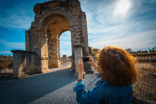Tourist woman taking travel picture with camera of the Arch of Caparra, famous tetrapylum in The Roman city of Caparra, now permanently abandoned. Located in the north of Extremadura, Spain.