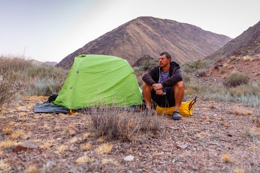 a man sits on a yellow bag near a tent in the mountains of Kyrgyzstan.