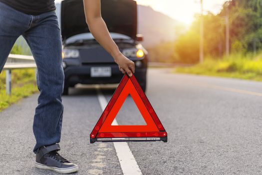 Man hold and put down the red triangle for warning have black color car parking  on road and oper car hood.