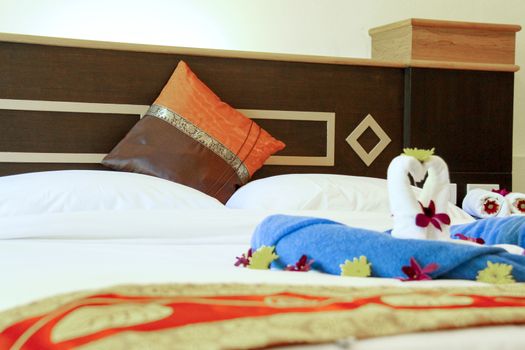 Two swans made from white towels are kissing on bed with yellow flower and orchid. White pillow and wood bed head.