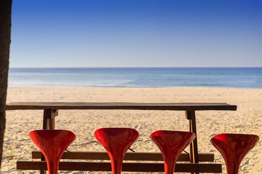 Holiday concept. 4 red bar chair with long wood table in fornt of beach and clear sky. With copy space for text or design.