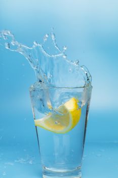 Lemon slice falling and dropping into a splashing glass with light blue background.