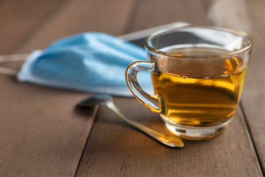 Close up hot tea cup on wood table with tea spoon, medical face mask and tablet . Soft focus and selective focus to tea cup. hot tea in glass with copy space for text or design. New normal concept.