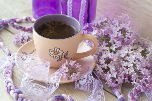 Violet cup of morning coffee or cappuccino and delicate purple, lilac flowers. Mother's day concept. Cozy breakfast