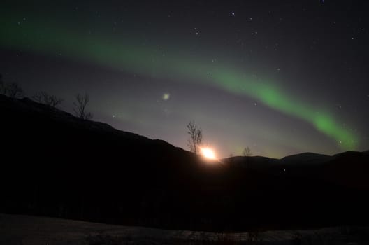 Aurora borealis pierce the night sky and the moon is rising up behind the mountain