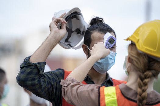 Factory woman worker in a face medical mask and safety dress used measures temperature at worker people standing on queue with a non-contact infrared thermometer.

