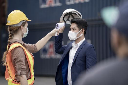 Factory woman worker in a face medical mask and safety dress used measures temperature at worker people standing on queue with a non-contact infrared thermometer. background of cargo container.
